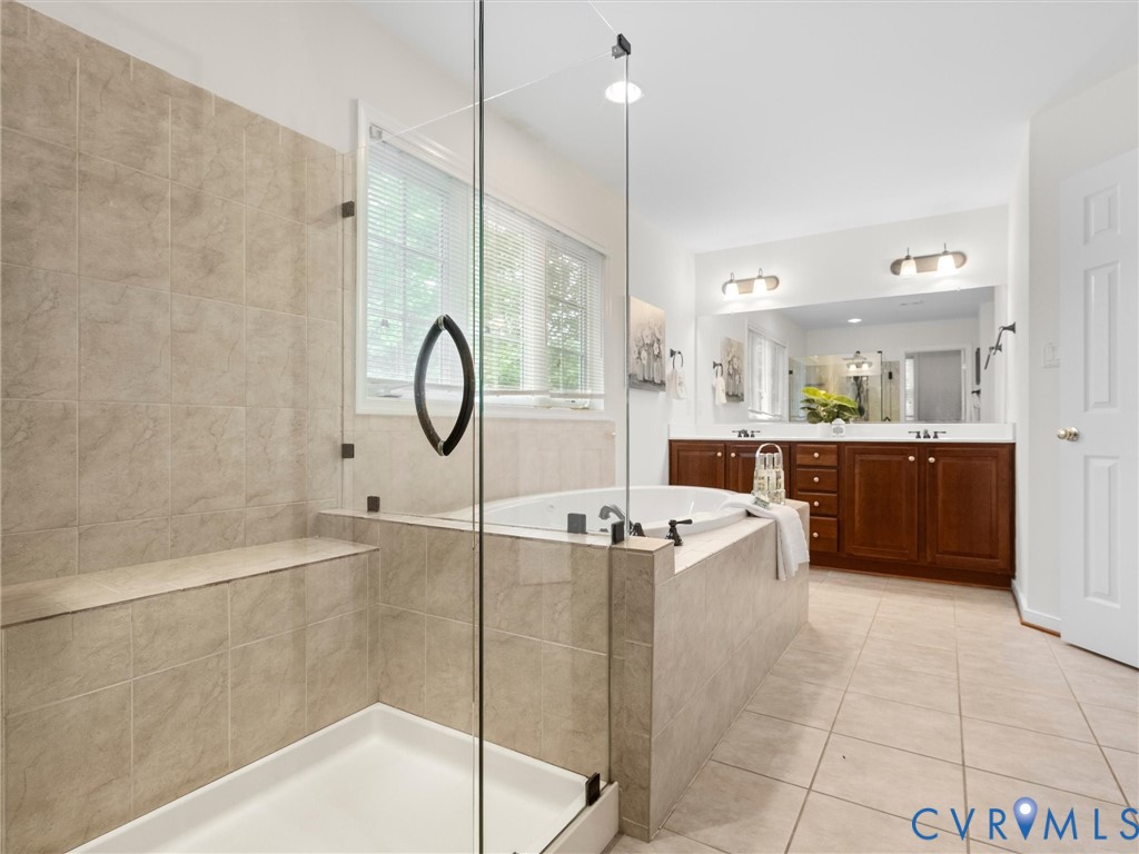 Spa-inspired primary bathroom featuring a frameless glass-enclosed shower with tile surround, a soaking tub positioned for relaxation, dual vanities with wood cabinetry and ample storage, polished nickel fixtures, and neutral tile flooring throughout