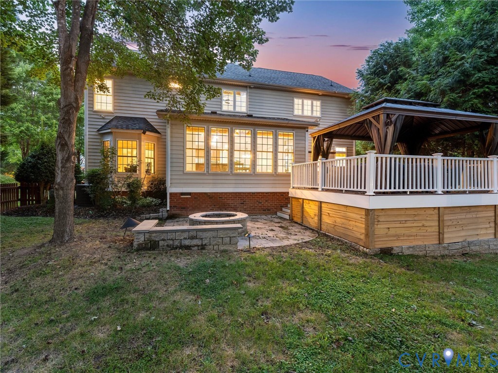 Rear exterior with siding, a bay window, a sunroom with multiple windows, a raised deck with white railings, and a built-in fire pit with a paver surround