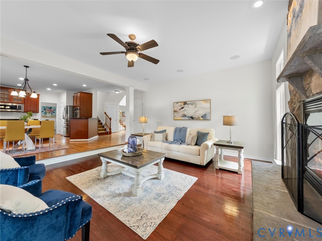 Hardwood flooring, stone fireplace, ceiling fan, recessed lighting, and kitchen with dark wood cabinetry