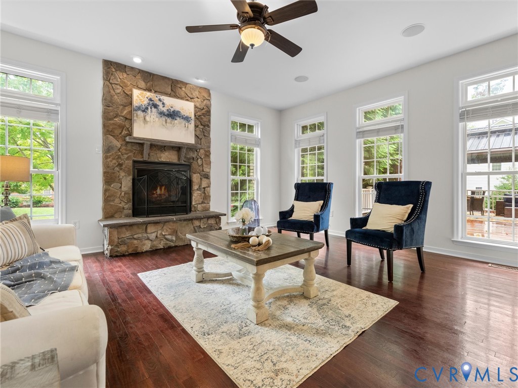 Stone-clad fireplace with a hearth, dark wood-finish flooring, multiple pane windows, ceiling fan with integrated lighting, and recessed ceiling lights