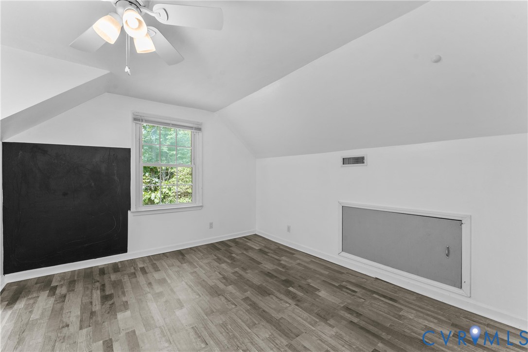 Sloped ceiling room featuring wood-finish flooring, a window with natural light, and a ceiling fan with integrated lighting