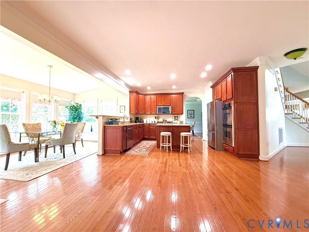Open-concept kitchen and dining area featuring warm wood-finish flooring, rich wood cabinetry, stainless steel appliances, and a central kitchen island with breakfast bar seating Open-concept kitchen and dining area featuring warm wood-finish flooring, rich wood cabinetry, stainless steel appliances, and a central kitchen island with breakfast bar seating