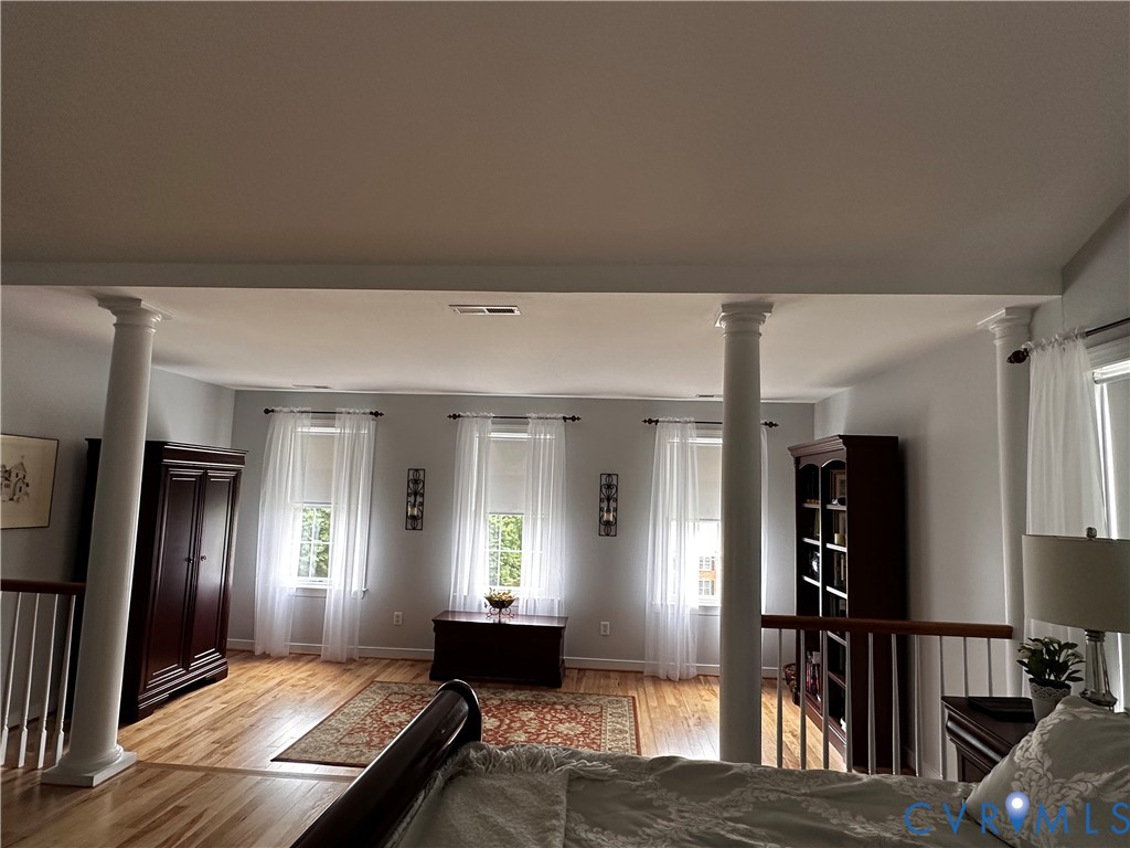 Light-filled open space featuring decorative columns, light wood-finish flooring, and multiple windows with sheer white drapery