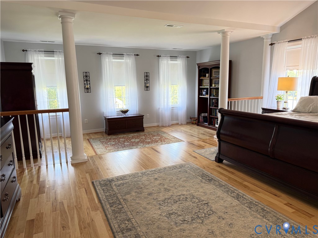 Spacious room featuring light wood-finish flooring, multiple windows with sheers, and architectural columns