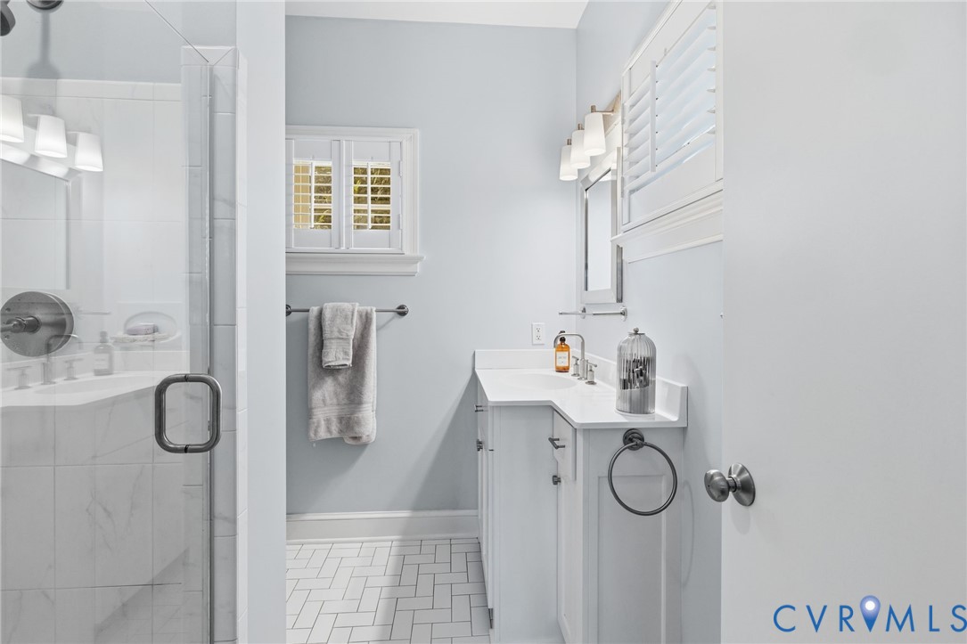 Bathroom featuring a glass-enclosed shower with white tiled walls, a single vanity with a white countertop and undermount sink, a wall-mounted mirror, and white plantation shutters on the window