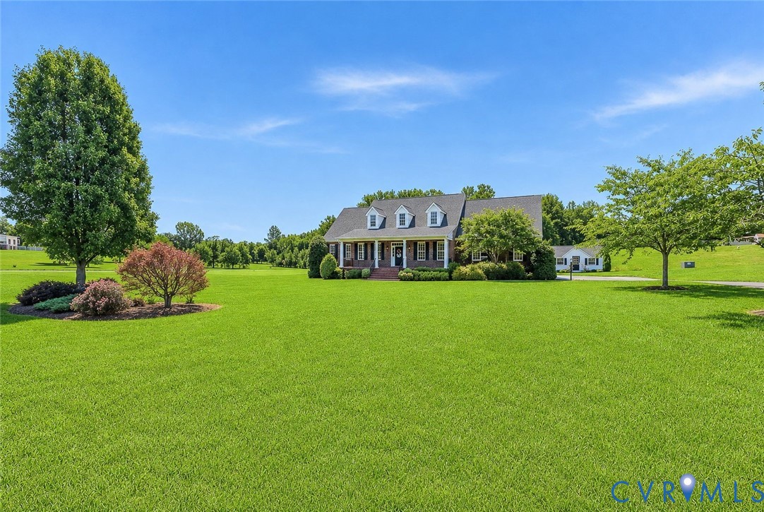 Expansive green lawn with mature trees and landscaping surrounding a brick residence featuring a covered front porch and dormer windows Expansive green lawn with mature trees and landscaping surrounding a brick residence featuring a covered front porch and dormer windows