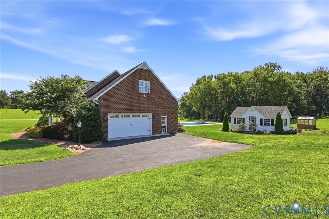 Brick facade residence featuring an attached two-car garage, expansive green lawn, separate white outbuilding, inground swimming pool, and a dedicated greenhouse structure