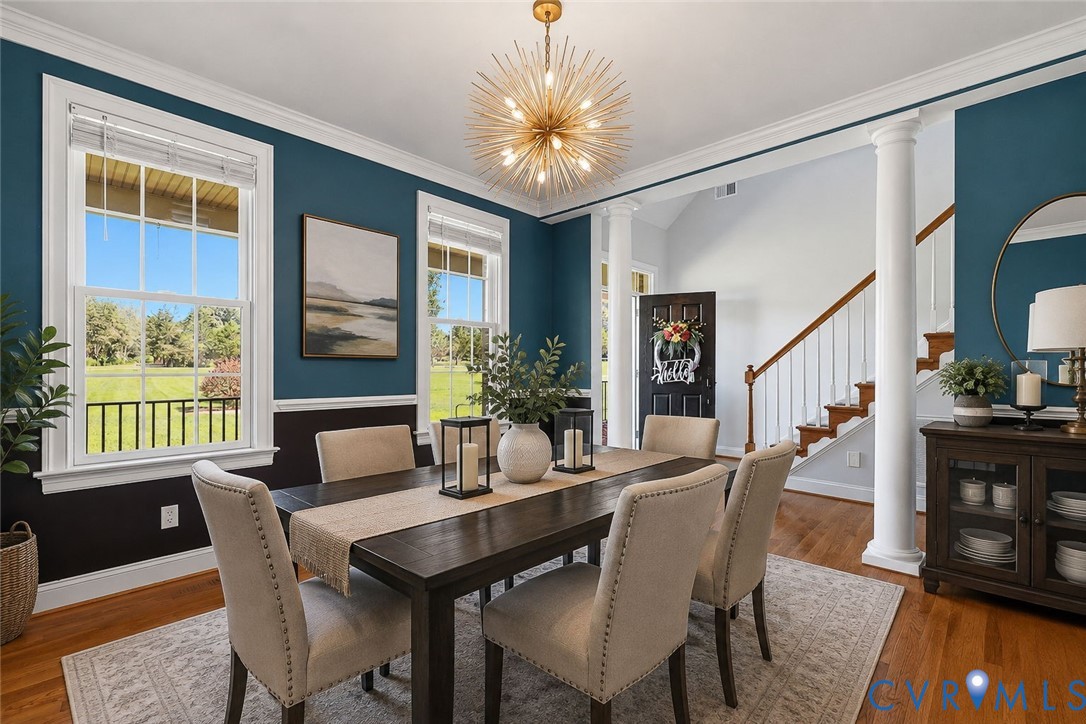 Formal dining area featuring hardwood flooring, a contemporary sputnik chandelier, two large windows, a decorative wainscoting, and an archway with columns leading to a staircase