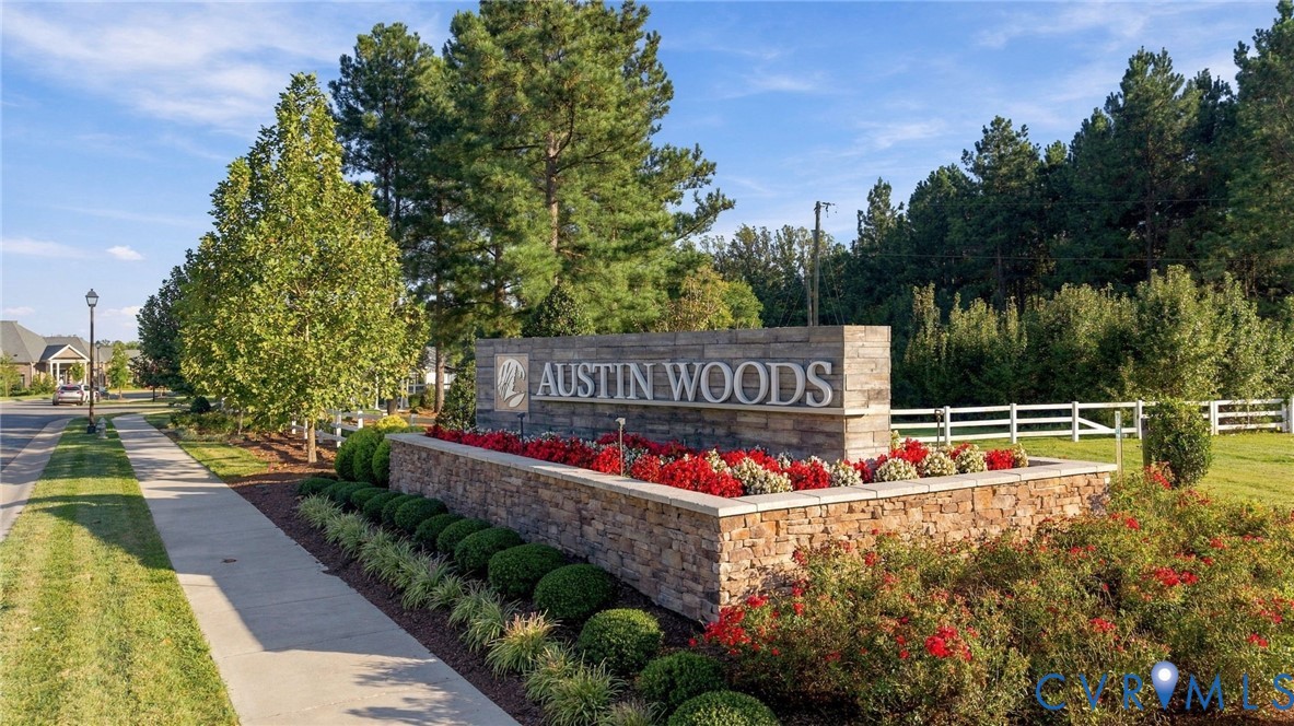 Stone-clad community entrance sign featuring "Austin Woods" lettering, decorative landscaping with red and green plantings, and a sidewalk leading into the community