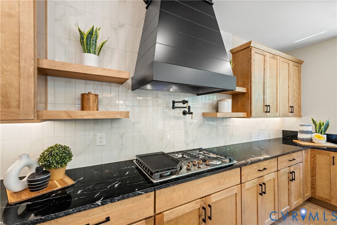Gourmet kitchen featuring a black range hood, gas cooktop, dark stone countertops, light wood cabinetry, and a white tile backsplash