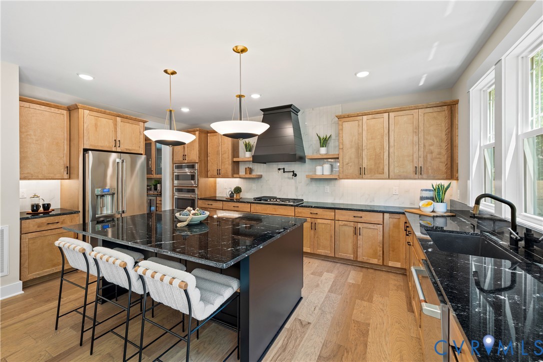Spacious kitchen featuring a large central island with a dark countertop, light wood-finish cabinetry, stainless steel appliances, a black range hood, and light wood-finish flooring