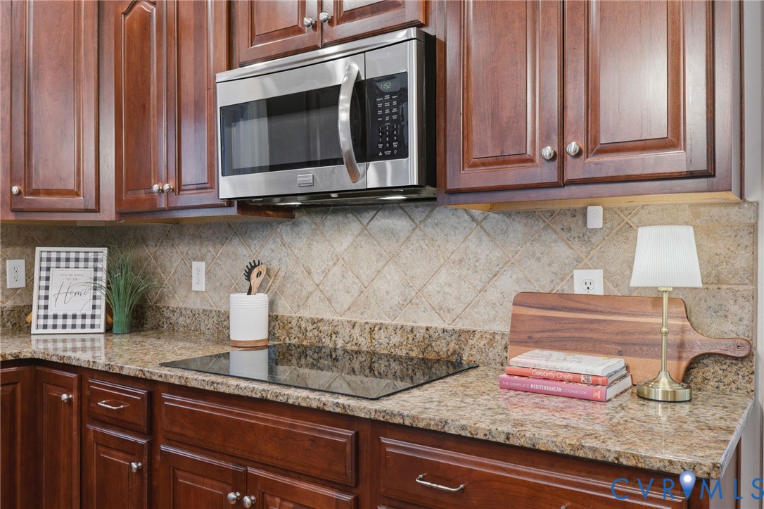Rich wood cabinetry with silver hardware, granite countertops, stainless steel microwave, black electric cooktop, and diamond-patterned tile backsplash
