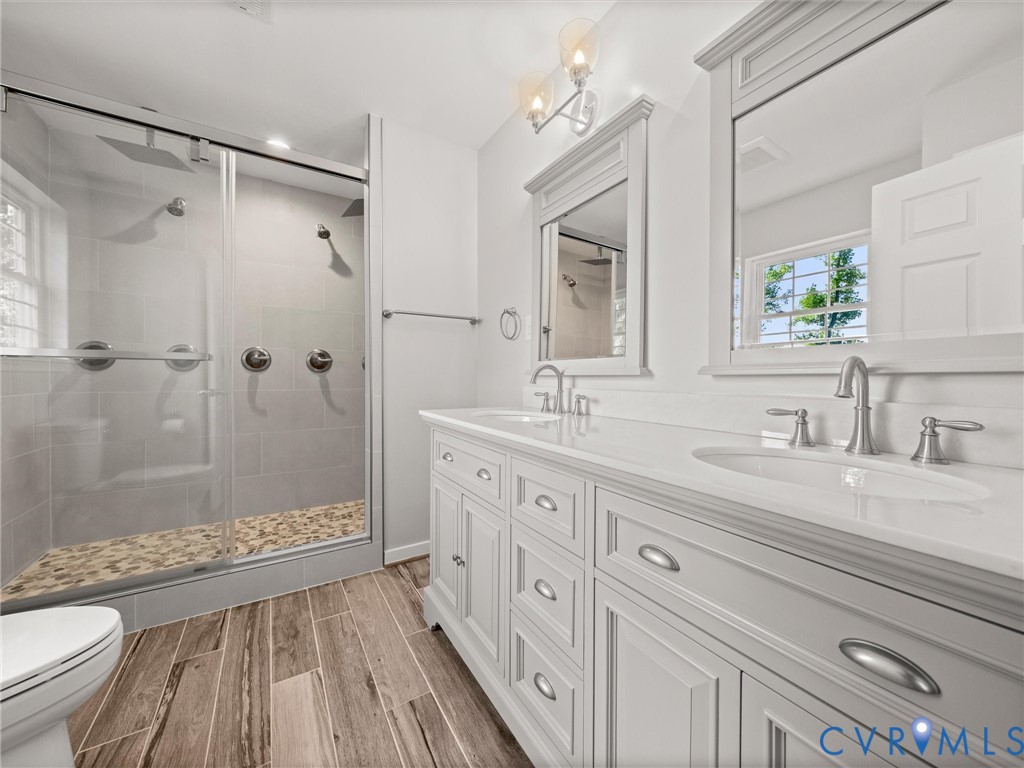 Dual vanity with undermount sinks, wood-finish flooring, glass-enclosed shower with pebble-tiled floor, and dual shower heads