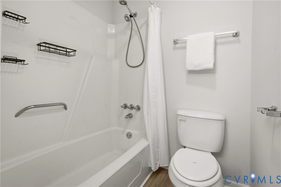 Clean and functional bathroom featuring a white porcelain toilet with elongated bowl, ceramic tile surround with grab bar accessibility, chrome shower fixtures with handheld showerhead, white subway tile walls, and chrome towel bar and shelving