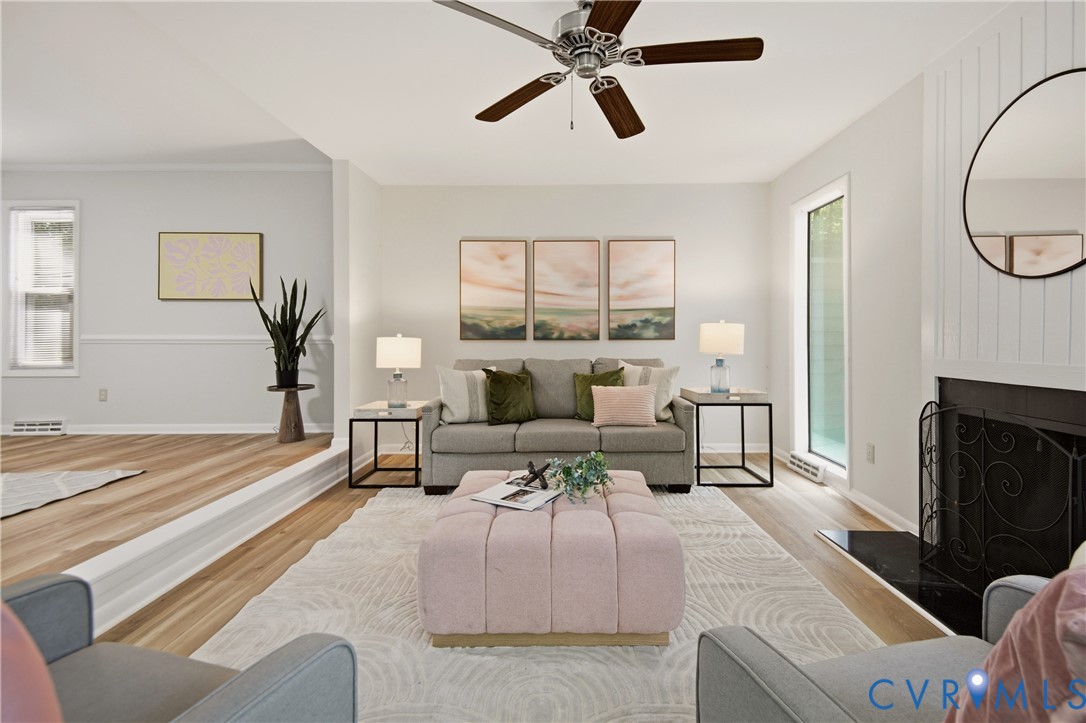 Living room featuring wood-finish flooring, a fireplace with a black hearth, a ceiling fan with dark wood-finish blades, and a built-in step separating distinct areas Living room featuring wood-finish flooring, a fireplace with a black hearth, a ceiling fan with dark wood-finish blades, and a built-in step separating distinct areas