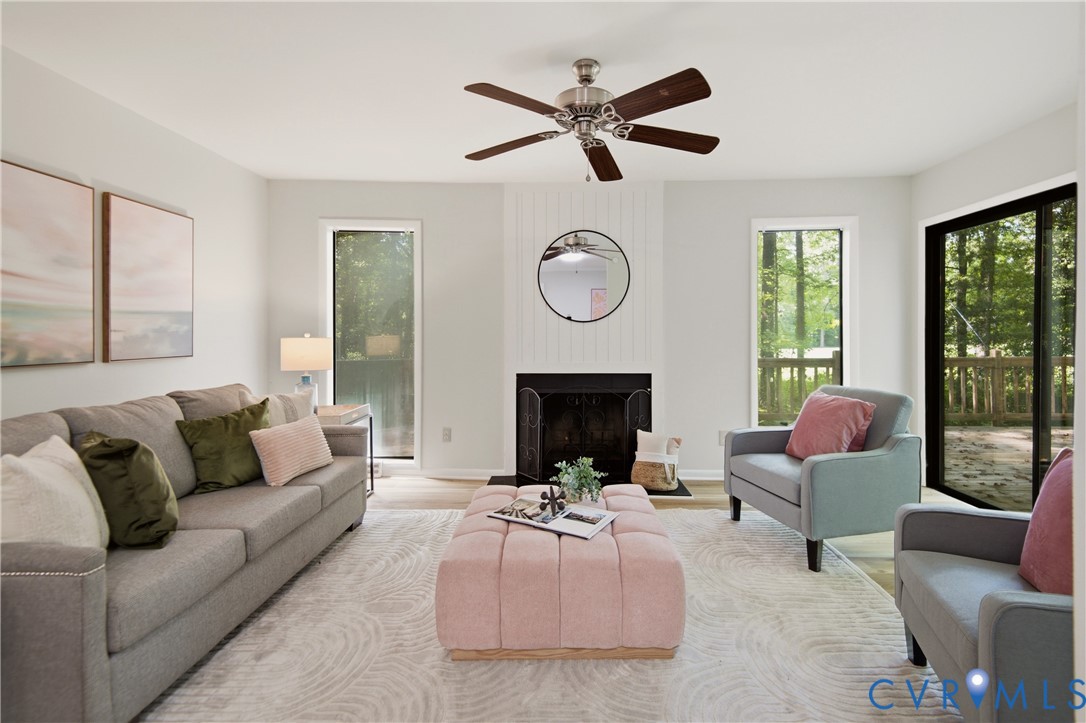 Bright, light-filled living room featuring expansive glass doors and windows framing natural outdoor scenery, a wood-finish ceiling fan with brushed metal accents, a fireplace with black surround, and neutral-toned wood-finish flooring throughout Bright, light-filled living room featuring expansive glass doors and windows framing natural outdoor scenery, a wood-finish ceiling fan with brushed metal accents, a fireplace with black surround, and neutral-toned wood-finish flooring throughout