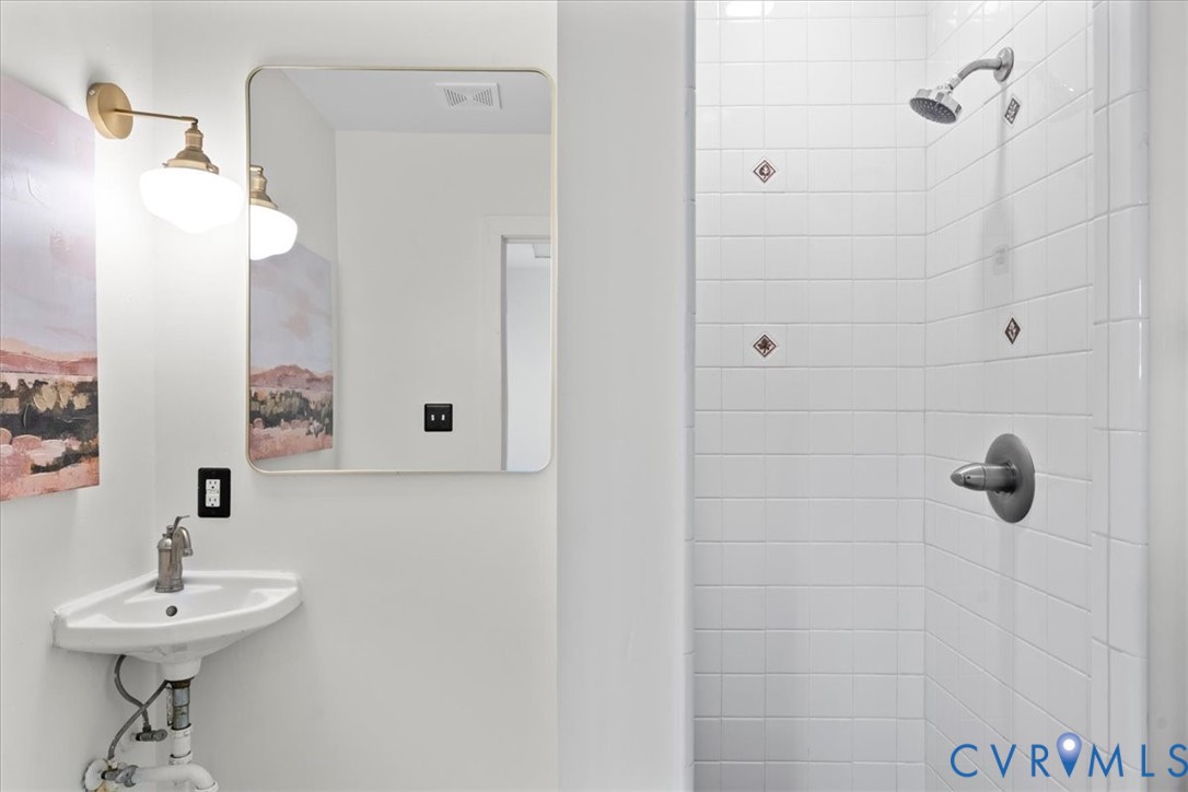White tile shower with decorative accent tiles, corner pedestal sink, and rectangular framed mirror with wall-mounted brass light fixture