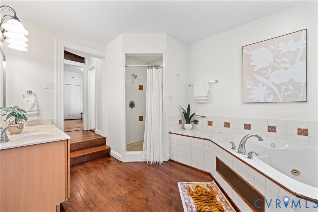 Hardwood flooring, built-in soaking tub, tiled shower enclosure, single vanity with integrated sink, and white wall tiling