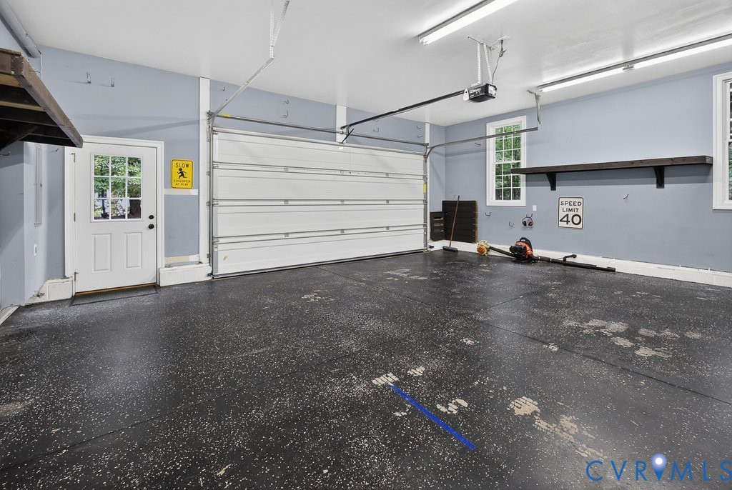 Spacious garage featuring a sectional overhead door, a pedestrian door with windowpanes, multiple windows, built-in shelving, and overhead lighting