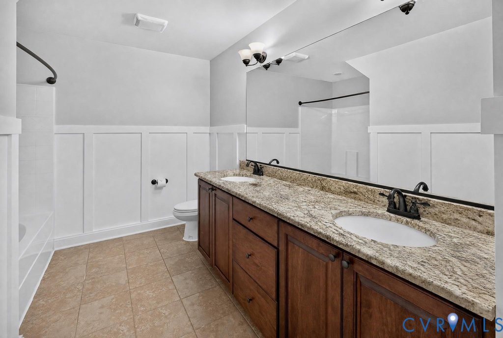 Double vanity with integrated sinks, granite countertop, rich wood-finish cabinetry, wainscoting, and large wall-mounted mirror
