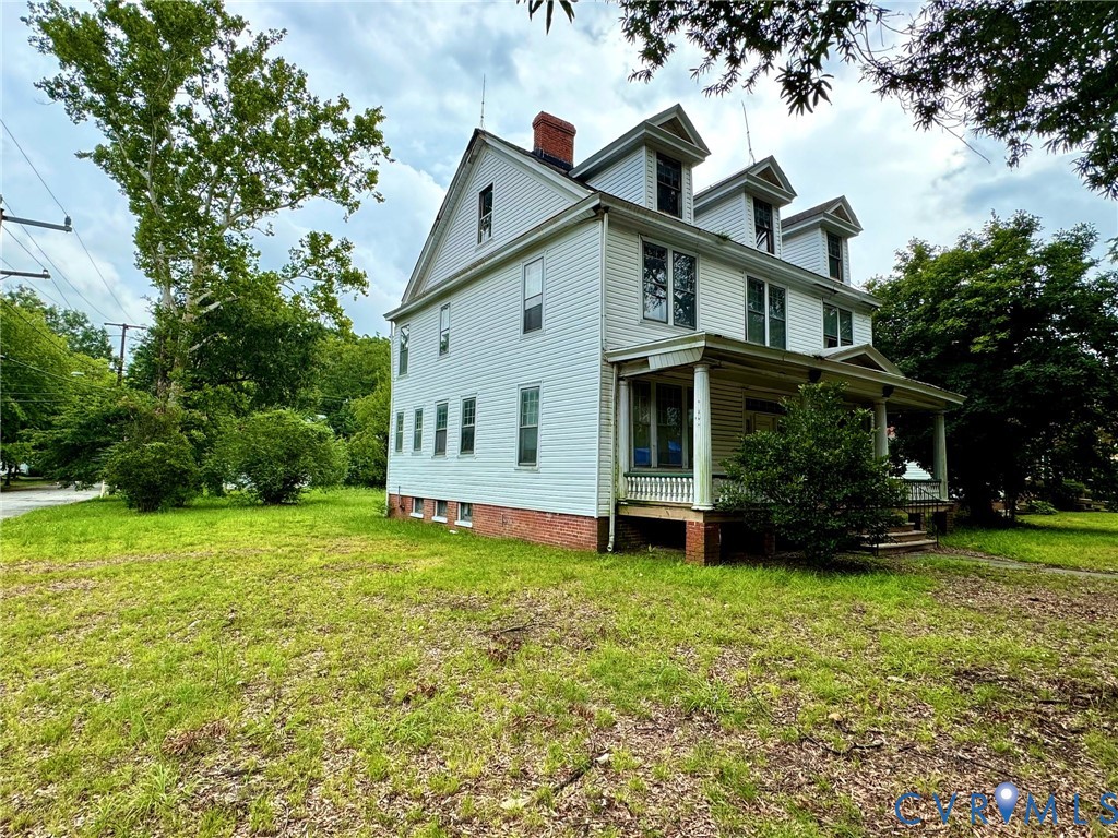 Multi-level residential structure featuring white siding, a covered porch, multiple dormer windows, a prominent brick chimney, and a surrounding lawn area with mature trees