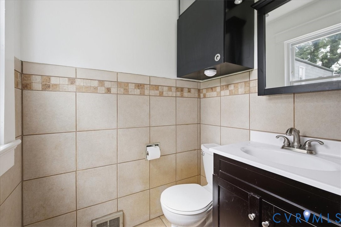Tile wainscoting with a decorative accent band, dark wood-finish vanity with an integrated sink, wall-mounted mirror, and an overhead storage cabinet