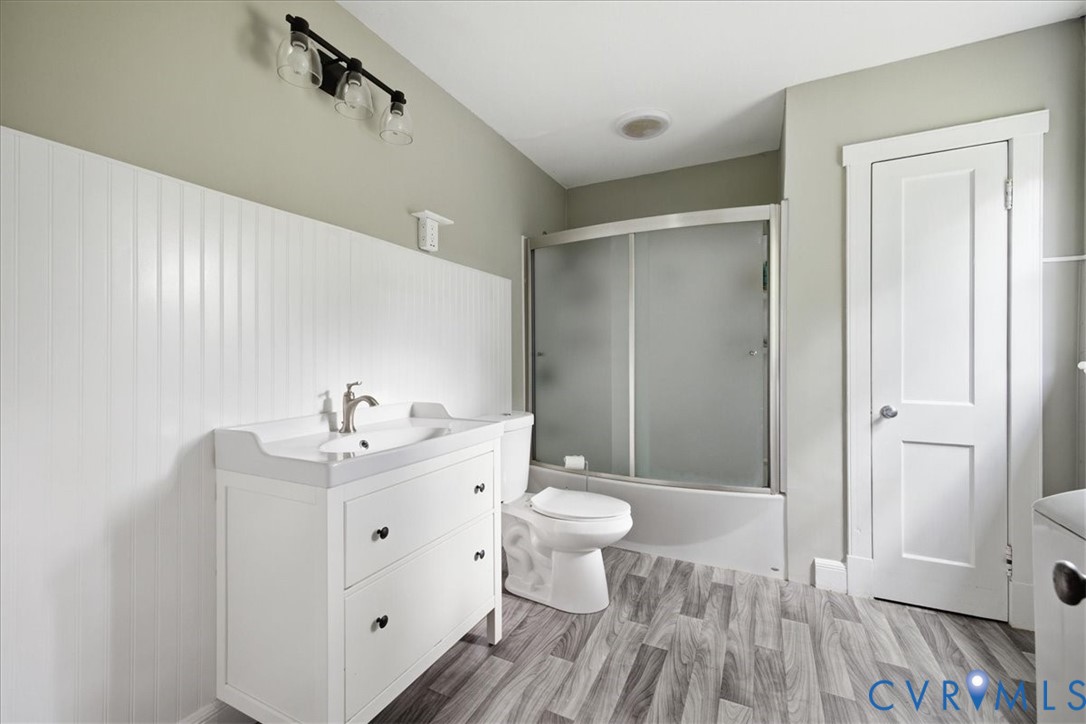 Bathroom featuring a white vanity with integrated sink, wood-finish flooring, a shower-tub combination with sliding frosted glass doors, and vertical paneling