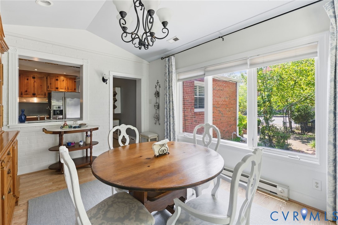Vaulted ceiling with an ornate chandelier, large windows with roller shades, wood-finish flooring, and a pass-through opening to the kitchen featuring wood cabinetry and stainless steel appliances