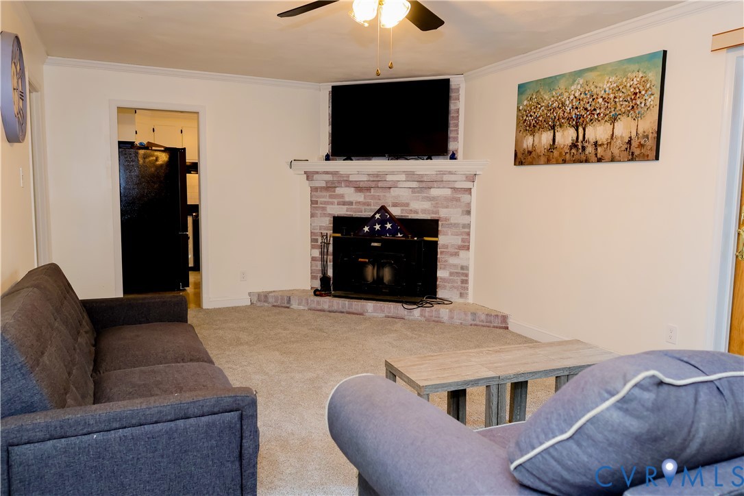 Recessed fireplace with brick surround and hearth, crown molding, ceiling fan with integrated lighting, and carpeted flooring