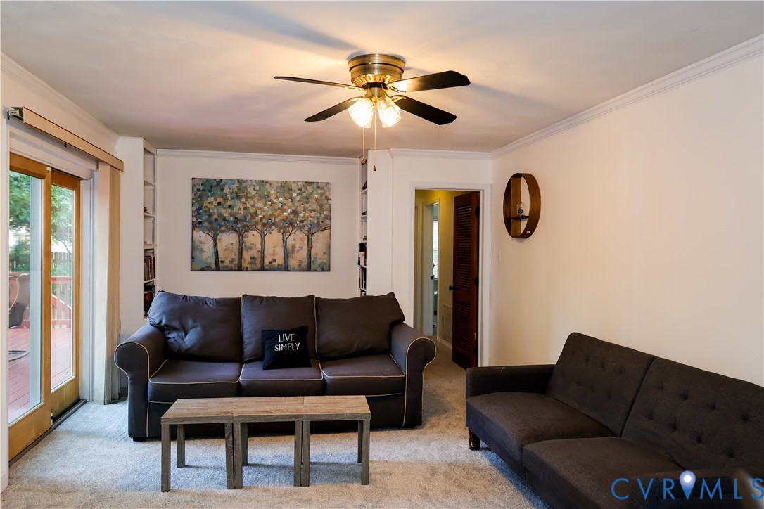 Recessed built-in shelving, ceiling fan w lighting, sliding glass door with wood-finish frame leads to massive deck, crown molding, and carpeted flooring.