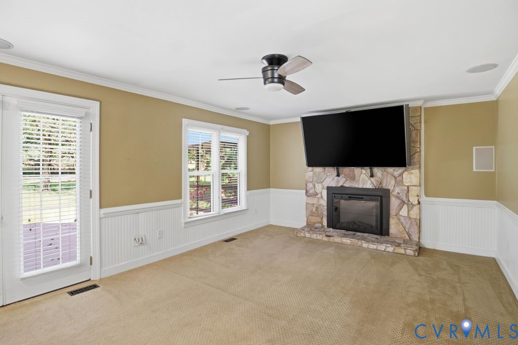 Stone fireplace with a black firebox, ceiling fan with wood-finish blades, white wainscoting, and crown molding