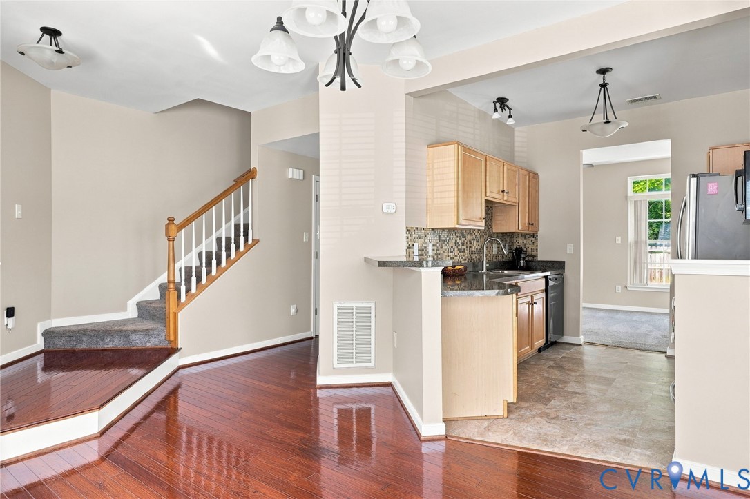 Hardwood-finish flooring, staircase with carpeted treads and wood railing, kitchen with wood-finish cabinetry, granite-style countertops, and a tiled backsplash Hardwood-finish flooring, staircase with carpeted treads and wood railing, kitchen with wood-finish cabinetry, granite-style countertops, and a tiled backsplash