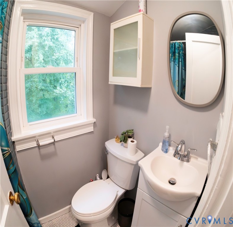 Single-hung window with white trim, wall-mounted storage cabinet with frosted glass panels, oval mirror with a gray frame, white pedestal sink with chrome faucet, and white hexagonal tile flooring