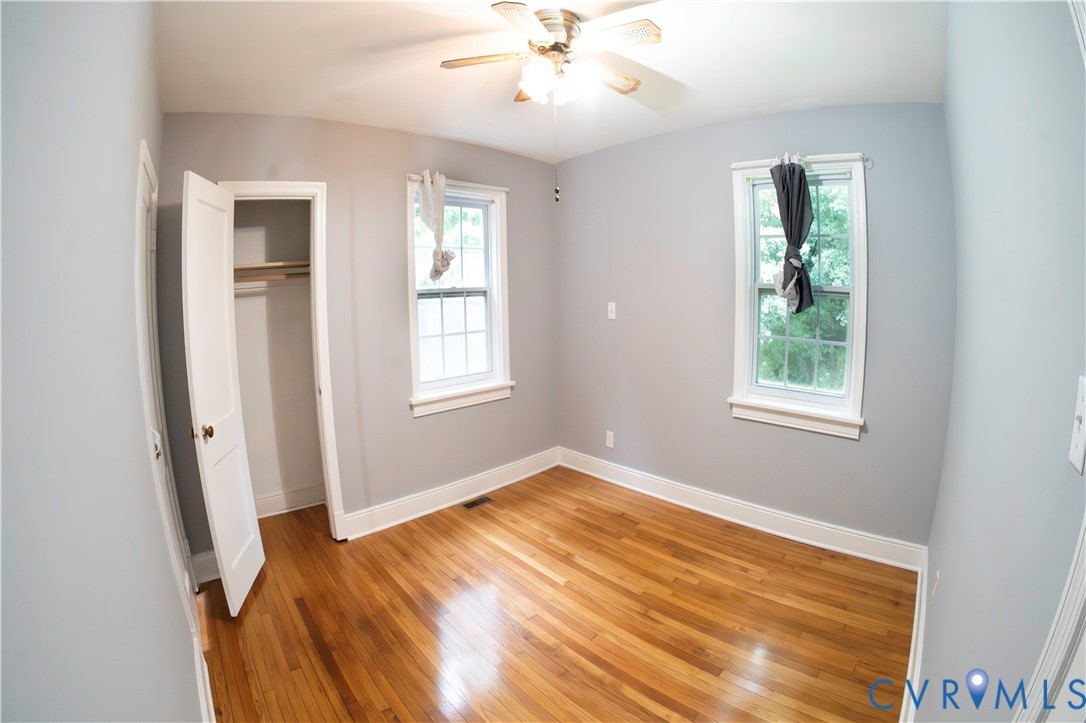Hardwood flooring, light gray wall finish, two windows with white trim, built-in closet with shelving, and a ceiling fan