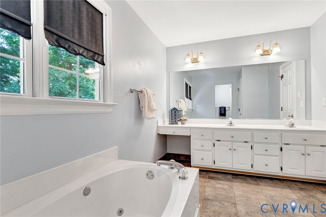 Soaking tub with integrated jets, dual vanity with white cabinetry, light-toned countertops, and expansive wall mirror