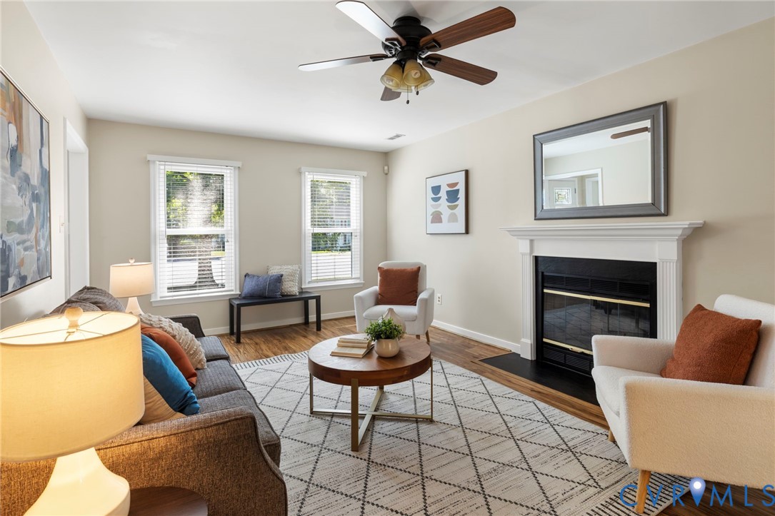 Living space featuring wood-finish flooring, two large windows with blinds, a ceiling fan, and a fireplace with a white mantel and black hearth Living space featuring wood-finish flooring, two large windows with blinds, a ceiling fan, and a fireplace with a white mantel and black hearth