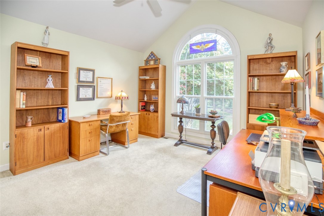 Second 1st floor bedroom or office suite featuring multiple wood cabinetry units with shelving and storage, abundant natural light from an arched transom window, wood-finish desk surfaces, ceiling fan, and neutral carpeting throughout the expansive room