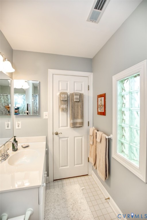 Bright, galley-style bathroom featuring a ceramic tile floor, white six-panel door with frosted glass inset, dual vanity with integrated sink and faucet, recessed ceiling lighting, and a window with decorative glass panes providing natural light