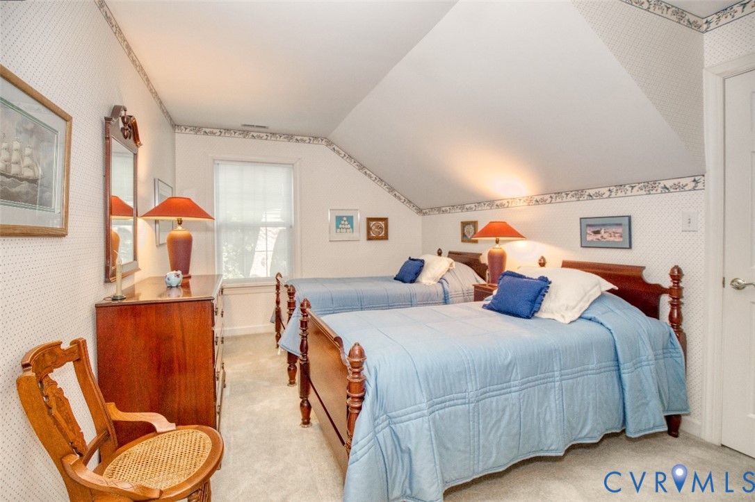 Charming second-floor bedroom featuring sloped vaulted ceilings with decorative trim, two twin beds with wooden posts, natural light from a front-facing window, warm wood-finish dresser, and coordinating table lamps with orange shades