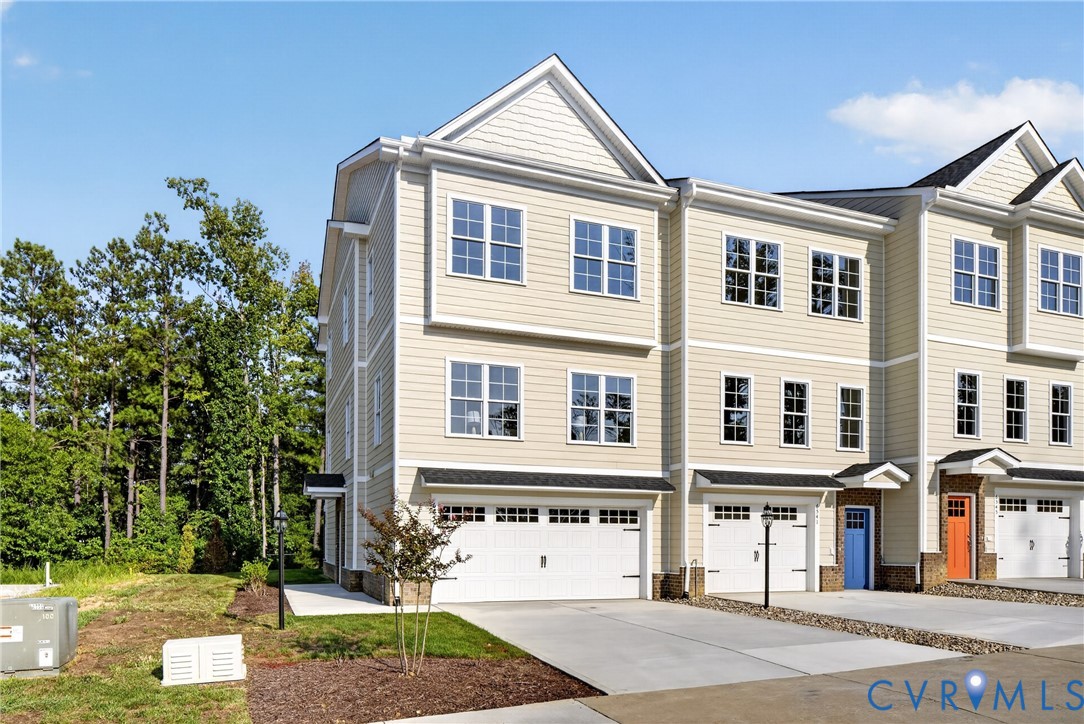 Multi-level townhome featuring light-colored siding, multiple windows, an attached garage, a paved driveway, and mature landscaping