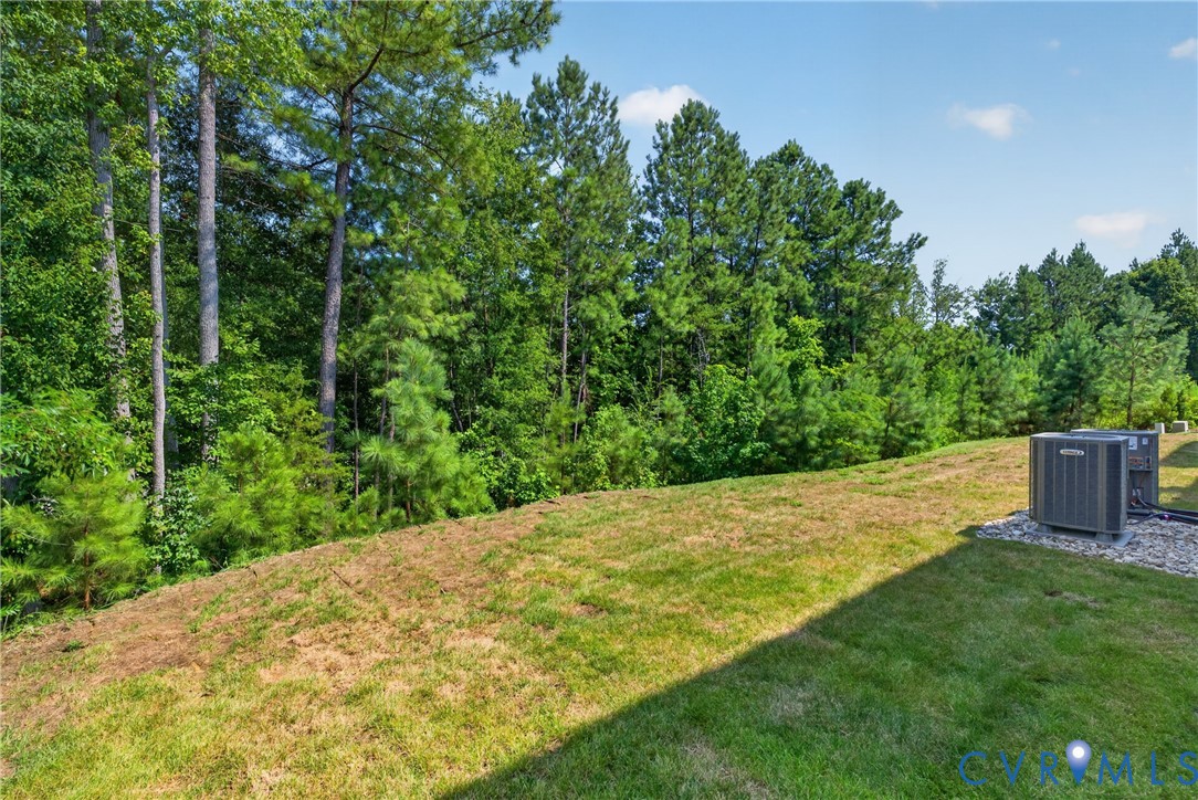 Expansive sloped lawn with a dense tree line backdrop, featuring two outdoor HVAC units on a gravel pad