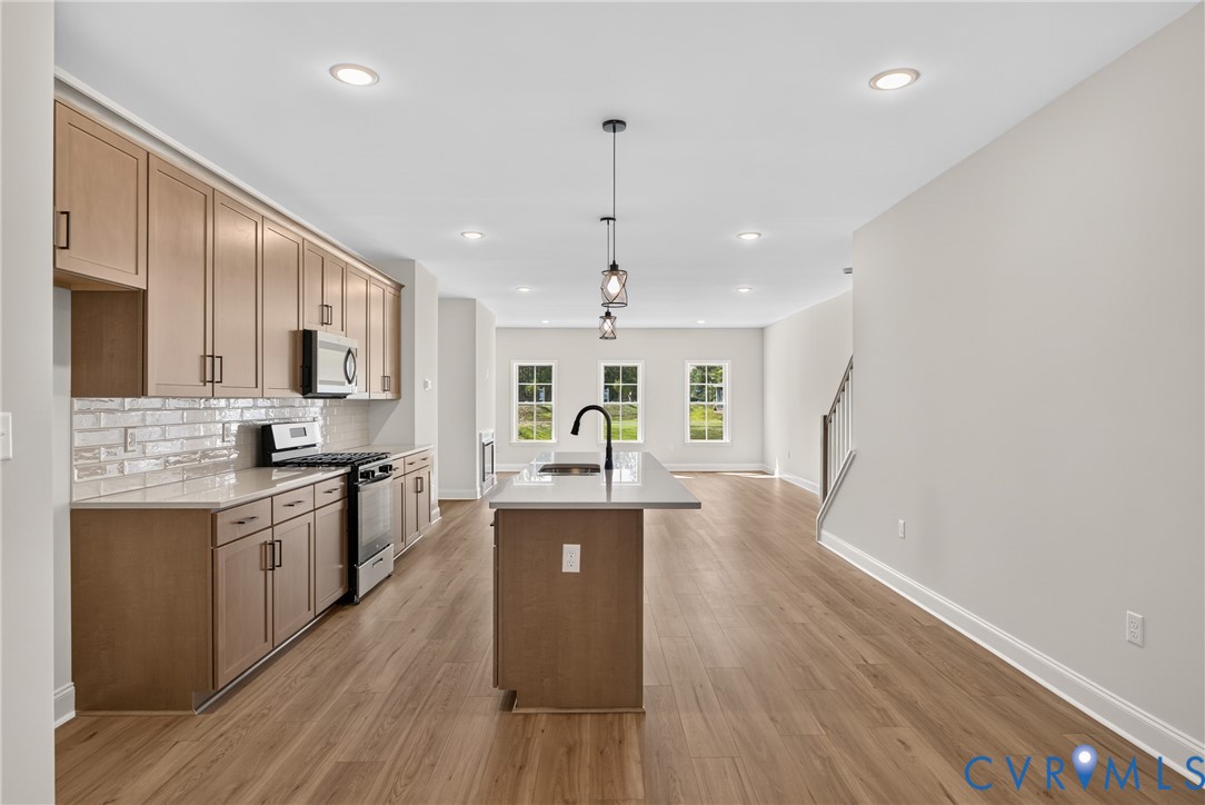Kitchen with wood-finish flooring, light-toned cabinetry, stainless steel appliances, a white subway tile backsplash, and a central island with a sink and pendant lighting
