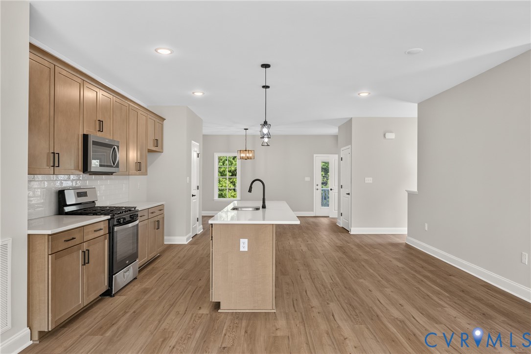 Kitchen featuring light wood-finish cabinetry, white subway tile backsplash, stainless steel appliances, and a central island with a sink and pendant lighting
