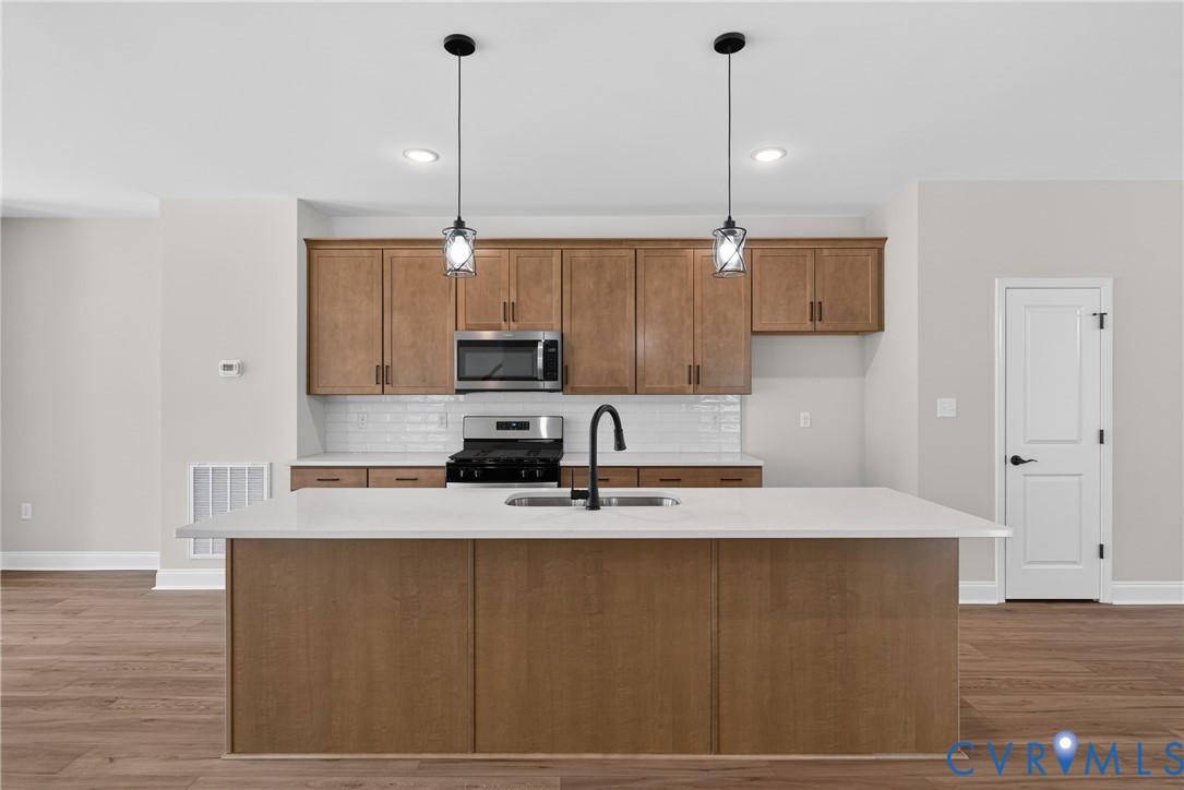Kitchen featuring wood-finish cabinetry, stainless steel appliances, a white subway tile backsplash, and an island with a white countertop and double sink