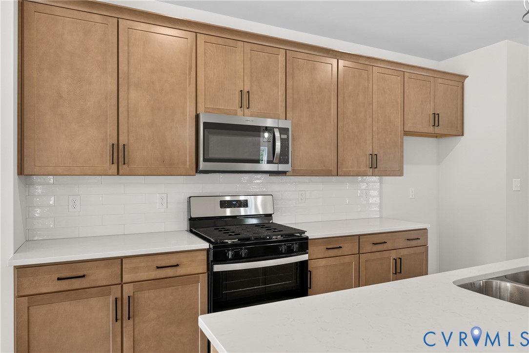 Wood-finish cabinetry, stainless steel microwave, white subway tile backsplash, white countertops, and stainless steel gas range