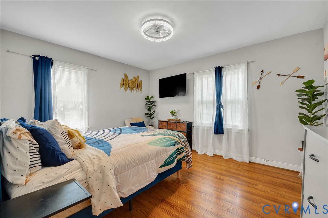Bright, spacious bedroom featuring hardwood flooring, dual glass doors with sheer and blue drapery, wall-mounted flat-screen television, ceiling fan with integrated lighting, and ample natural light from multiple windows