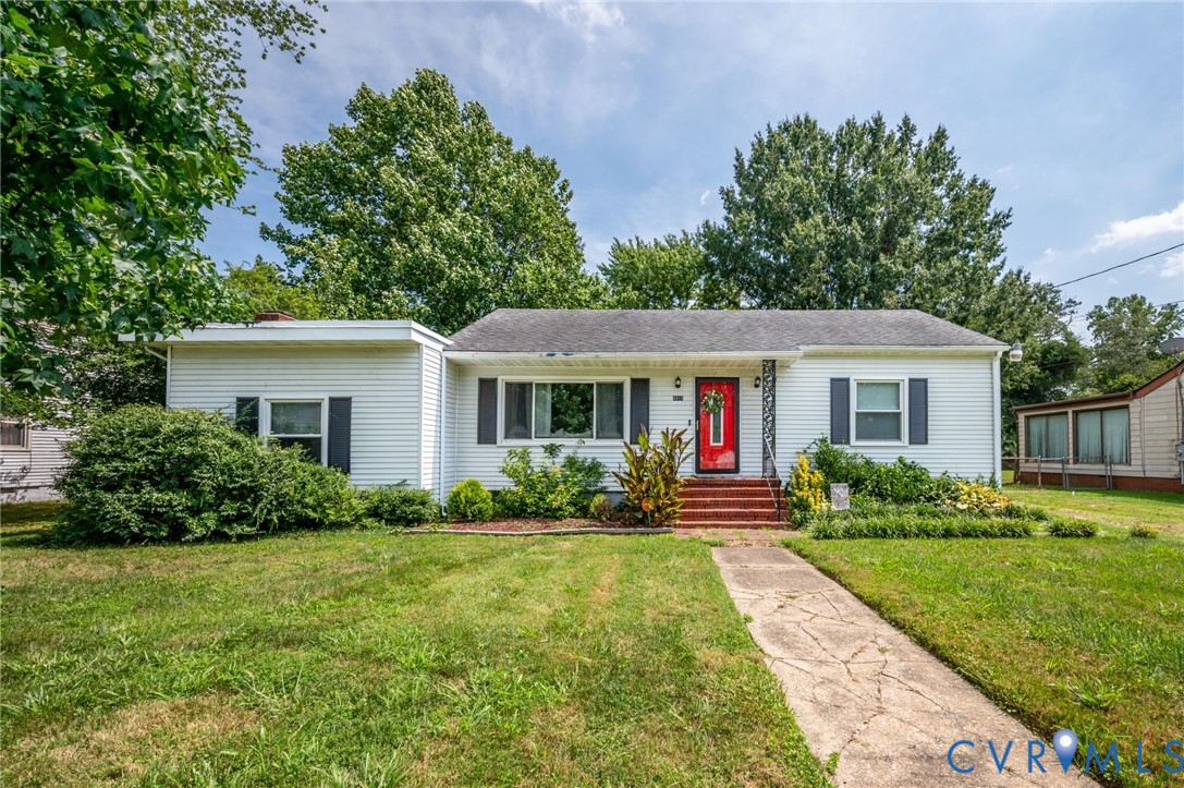 White siding exterior with dark shutters, red entry door, established landscaping, and a concrete pathway White siding exterior with dark shutters, red entry door, established landscaping, and a concrete pathway