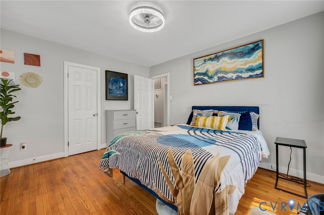 Bright secondary bedroom featuring hardwood flooring, a queen-sized bed frame with blue and gold upholstered headboard, large windows providing natural light, and multiple closet access points