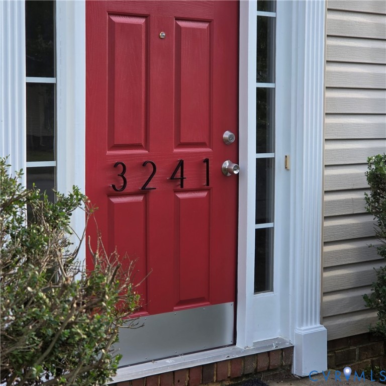 Red paneled entry door with sidelight windows, polished chrome hardware, and brick foundation