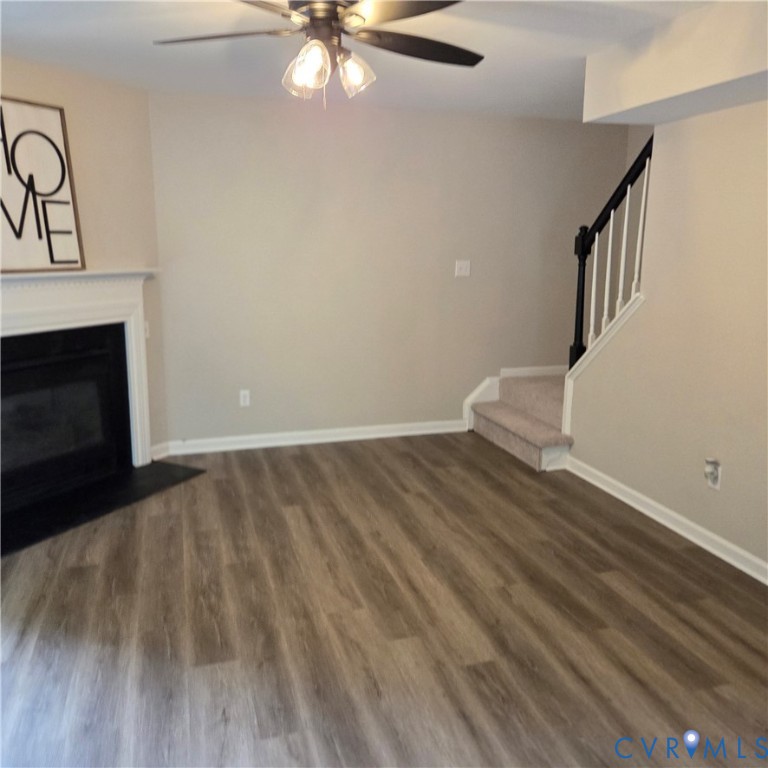 Wood-finish flooring, fireplace with a white mantel, ceiling fan with integrated lighting, neutral wall tones, and a carpeted staircase